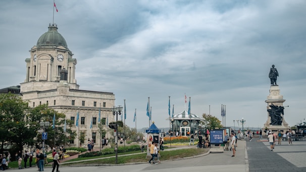 A historic building with a clock tower stands prominently, surrounded by people walking and engaging in leisure activities. A large statue is visible in the background, and flags are waving around a small pavilion amid flowerbeds.