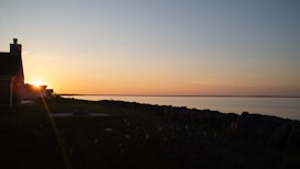 The image captures a serene sunset over a calm body of water. The horizon is lit with hues of orange, pink, and blue as the sun sets. A dark silhouette of a house is visible on the left side of the image, with a rocky shoreline stretching out towards the right. The peaceful setting evokes feelings of tranquility and calmness.