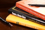 A stack of books is arranged on a wooden surface. The books include 'Aristotle's Nicomachean Ethics', 'Think & Grow Rich' by Napoleon Hill, and 'The Energy Bus'. Additionally, there are two fountain pens placed around the stack.