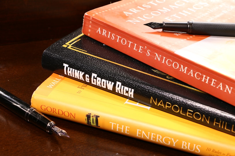 A stack of books is arranged on a wooden surface. The books include 'Aristotle's Nicomachean Ethics', 'Think & Grow Rich' by Napoleon Hill, and 'The Energy Bus'. Additionally, there are two fountain pens placed around the stack.