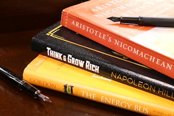 A stack of books is arranged on a wooden surface. The books include 'Aristotle's Nicomachean Ethics', 'Think & Grow Rich' by Napoleon Hill, and 'The Energy Bus'. Additionally, there are two fountain pens placed around the stack.
