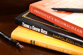 A stack of books is arranged on a wooden surface. The books include 'Aristotle's Nicomachean Ethics', 'Think & Grow Rich' by Napoleon Hill, and 'The Energy Bus'. Additionally, there are two fountain pens placed around the stack.