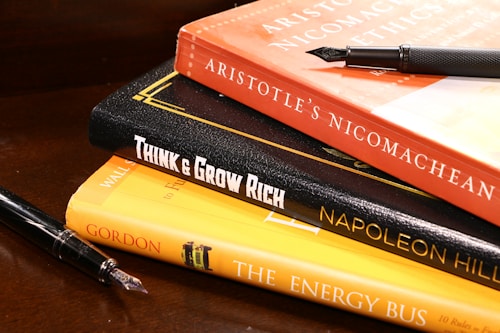 A stack of books is arranged on a wooden surface. The books include 'Aristotle's Nicomachean Ethics', 'Think & Grow Rich' by Napoleon Hill, and 'The Energy Bus'. Additionally, there are two fountain pens placed around the stack.