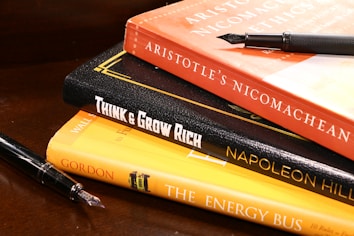 A stack of books is arranged on a wooden surface. The books include 'Aristotle's Nicomachean Ethics', 'Think & Grow Rich' by Napoleon Hill, and 'The Energy Bus'. Additionally, there are two fountain pens placed around the stack.