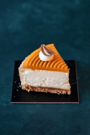 A slice of cheesecake with a crumbly base, creamy middle layer, and an orange-tinted top layer with decorative grooves. A small dollop of white cream topped with a toasted, brown-tipped garnish sits on top.