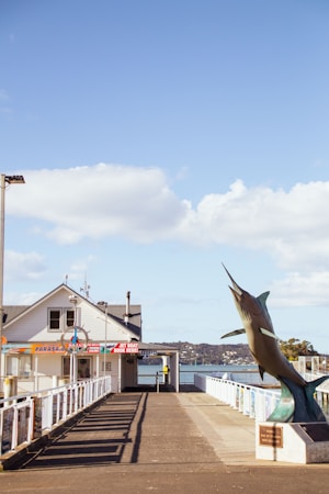 A pier leading to a building with various signs offering parasailing and jet boat bookings. To the right, a large statue of a marlin fish stands prominently. The background features a serene body of water and a clear sky with some scattered clouds.