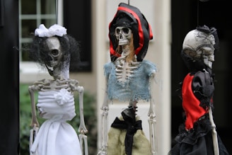 Three skeleton figures dressed in various pieces of fabric, including white, black, red, and blue, with varying textures. The central figure wears a hat made of red and black material, and each skeleton has distinctive clothing and features, giving them a spooky appearance.