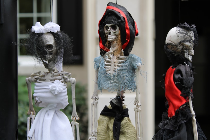 Three skeleton figures dressed in various pieces of fabric, including white, black, red, and blue, with varying textures. The central figure wears a hat made of red and black material, and each skeleton has distinctive clothing and features, giving them a spooky appearance.