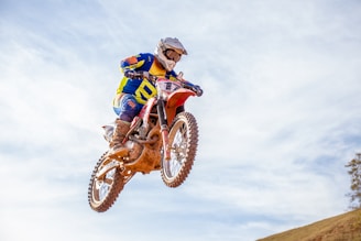A motorcyclist wearing a helmet and colorful gear performs an impressive jump on a dirt bike against a clear blue sky. The bike is airborne, kicking up dust from the rear tire, and the rider is shown in mid-air with a dynamic posture.