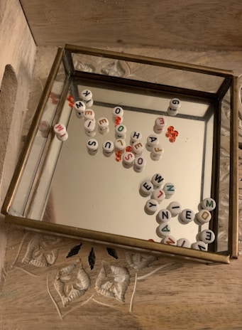 A glass tray with a mirrored base contains scattered round white beads with alphabet letters. There are also small red beads. The tray is placed on a wooden surface with carved patterns.