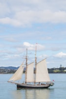 A large sailboat or schooner with multiple masts and white sails is navigating through calm waters. The vessel is positioned slightly to the right, with a spacious deck visible. In the background, there are hills and a coastline, under a mostly cloudy sky with patches of blue.