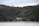 A serene landscape featuring a secluded white house nestled amidst dense green forests. The house is located near a tranquil shoreline with a wooden dock extending into calm waters, set against a backdrop of tall trees.