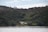 A serene landscape featuring a secluded white house nestled amidst dense green forests. The house is located near a tranquil shoreline with a wooden dock extending into calm waters, set against a backdrop of tall trees.