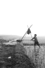 A person is using a manual water pump, standing in a field with young plants. The landscape is rural, with visible irrigation channels and an overcast or foggy sky in the background.