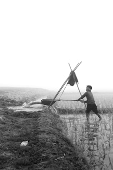 A person is using a manual water pump, standing in a field with young plants. The landscape is rural, with visible irrigation channels and an overcast or foggy sky in the background.