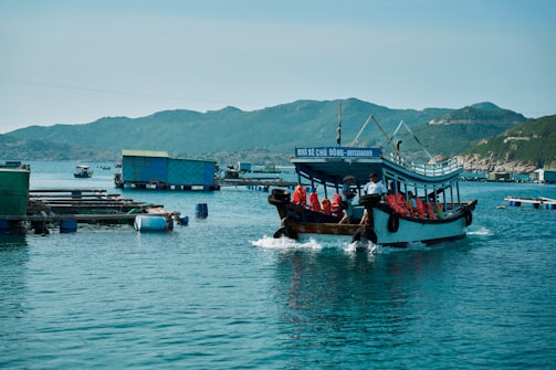 A wooden boat carrying several people wearing orange life jackets travels through a calm body of water. The boat is painted blue and has a sign with Vietnamese text. In the background, there are floating structures and green hills under a clear blue sky.