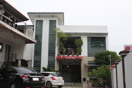 A modern building with two vertical glass sections flanking the entrance, which has a red sign with text and some potted plants. The facade includes green-tinted windows and foliage, with a mixture of rectangular architectural elements. The structure is surrounded by parked cars and other buildings, contributing to an urban atmosphere.