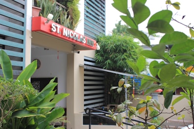 An exterior view of a building entrance featuring a sign with white lettering on a red background reading 'St. Nicolas Inn.' The entrance is surrounded by lush green foliage, potted plants, and shrubs, with modern architectural elements such as horizontal slats. The scene conveys a sense of inviting greenery and tranquility.