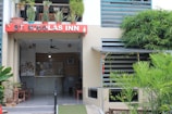 A building entrance with a red sign labeled 'St. Nicolas Inn' is adorned with various potted plants on a balcony above. Below the sign is a reception area with a fan, a small light, and some seating arrangements. The surroundings feature lush green plants and a modern exterior design.
