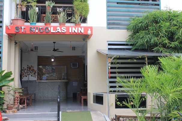 A building entrance with a red sign labeled 'St. Nicolas Inn' is adorned with various potted plants on a balcony above. Below the sign is a reception area with a fan, a small light, and some seating arrangements. The surroundings feature lush green plants and a modern exterior design.