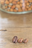 Three coffee beans are placed on a wooden surface with a blurred background showing a glass container filled with various nuts.
