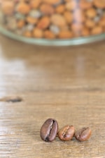 Three coffee beans are placed on a wooden surface with a blurred background showing a glass container filled with various nuts.