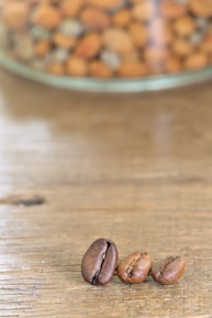 Three coffee beans are placed on a wooden surface with a blurred background showing a glass container filled with various nuts.