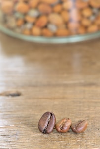 Three coffee beans are placed on a wooden surface with a blurred background showing a glass container filled with various nuts.