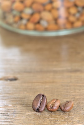 Three coffee beans are placed on a wooden surface with a blurred background showing a glass container filled with various nuts.