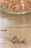 Three coffee beans are placed on a wooden surface with a blurred background showing a glass container filled with various nuts.