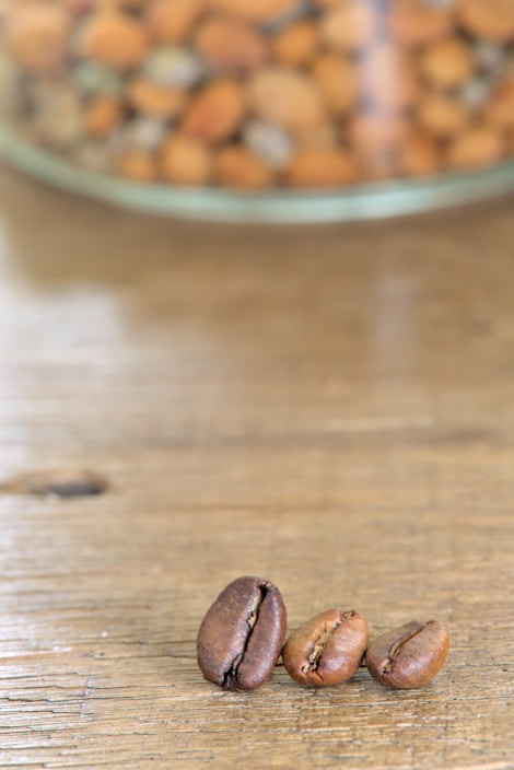 Three coffee beans are placed on a wooden surface with a blurred background showing a glass container filled with various nuts.