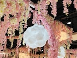 A ceiling decorated with cascading pink and cream artificial flowers. Large, cloud-like pendant lights hang among the floral arrangements, casting a soft glow. The black ceiling contrasts with the colorful, delicate blooms.