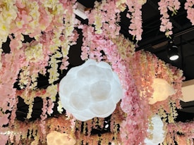 A ceiling decorated with cascading pink and cream artificial flowers. Large, cloud-like pendant lights hang among the floral arrangements, casting a soft glow. The black ceiling contrasts with the colorful, delicate blooms.