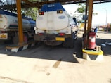 A large industrial fuel tanker truck is parked at a loading station. The truck is positioned under a roofed structure with supporting pillars. There are two visible tanks labeled 'Industri' and 'Solar'. Nearby, a fire extinguisher is mounted on a platform. There are traffic cones and safety signs present, suggesting a controlled environment. Some people are present in the background, wearing work-related clothing.