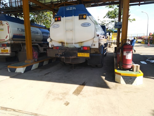 A large industrial fuel tanker truck is parked at a loading station. The truck is positioned under a roofed structure with supporting pillars. There are two visible tanks labeled 'Industri' and 'Solar'. Nearby, a fire extinguisher is mounted on a platform. There are traffic cones and safety signs present, suggesting a controlled environment. Some people are present in the background, wearing work-related clothing.