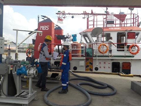 Two workers in protective gear and helmets are standing on a dock near machinery, engaged in discussion. A red and white boat is docked alongside, with several lifebuoys visible. Another person is seen walking on the boat's deck.