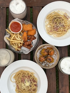 A wooden table features a variety of dishes, including plates of spaghetti with creamy sauce and bacon bits, a basket containing golden fried chicken, fries, and sausages, a serving of sushi-like rolls topped with sauce and herbs, and three glasses filled with creamy, frothy beverages.