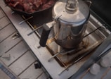 A metal coffee percolator is placed on a portable stove burner. Next to it, a cast iron skillet contains pieces of raw meat. The stove shows signs of usage with some stains and a bit of grit around the burner area. The scene suggests an outdoor or camping cooking setup with a rustic feel.