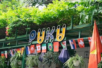 A colorful sign with the word 'PUKU' in bold letters is surrounded by lush green plants. Beneath it, there is a variety of small national flags hanging in a row. The flags include those of Canada, Switzerland, and several others, suggesting a multicultural or international theme. The background is filled with leafy vegetation and hanging potted plants.
