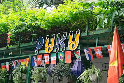A colorful sign with the word 'PUKU' in bold letters is surrounded by lush green plants. Beneath it, there is a variety of small national flags hanging in a row. The flags include those of Canada, Switzerland, and several others, suggesting a multicultural or international theme. The background is filled with leafy vegetation and hanging potted plants.