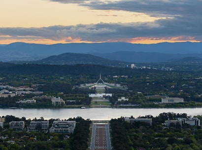 A picturesque view of a cityscape with a prominent road leading towards a large government building. Surrounding the central structure are lush green trees, and a wide river is in the foreground. The background features a range of hills under a cloudy sky, with the horizon tinted by sunset colors.