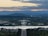 A picturesque view of a cityscape with a prominent road leading towards a large government building. Surrounding the central structure are lush green trees, and a wide river is in the foreground. The background features a range of hills under a cloudy sky, with the horizon tinted by sunset colors.
