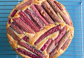 A round, baked cake featuring a topping of neatly arranged pieces of rhubarb, creating a pattern. The cake sits on a metal cooling rack placed on a light blue surface.