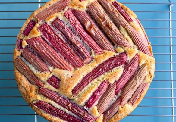 A round, baked cake featuring a topping of neatly arranged pieces of rhubarb, creating a pattern. The cake sits on a metal cooling rack placed on a light blue surface.