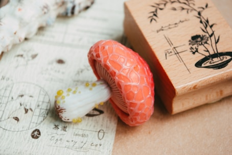 A detailed close-up of a decorative mushroom with a textured orange cap and a white stem adorned with yellow spots. The mushroom is positioned next to a wooden stamp featuring botanical illustrations and text, set against a background of light-colored paper with sketches and foreign writing.