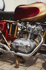 A close-up view of a detailed classic motorcycle engine, featuring polished metal and chrome components. The red and gold color scheme on the bodywork complements the intricate design of the mechanical parts.