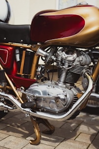 A close-up view of a detailed classic motorcycle engine, featuring polished metal and chrome components. The red and gold color scheme on the bodywork complements the intricate design of the mechanical parts.
