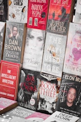 A display of various books with different genres, featuring both fiction and non-fiction works. The arrangement includes titles like 'Anxious People', 'Angels & Demons', 'The Book Thief', and 'The Way I Used To Be'. The covers are visually distinct with a mix of bold and muted colors, depicting elements and characters relevant to their stories.