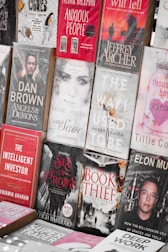 A display of various books with different genres, featuring both fiction and non-fiction works. The arrangement includes titles like 'Anxious People', 'Angels & Demons', 'The Book Thief', and 'The Way I Used To Be'. The covers are visually distinct with a mix of bold and muted colors, depicting elements and characters relevant to their stories.