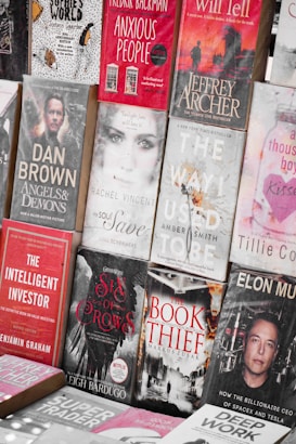 A display of various books with different genres, featuring both fiction and non-fiction works. The arrangement includes titles like 'Anxious People', 'Angels & Demons', 'The Book Thief', and 'The Way I Used To Be'. The covers are visually distinct with a mix of bold and muted colors, depicting elements and characters relevant to their stories.